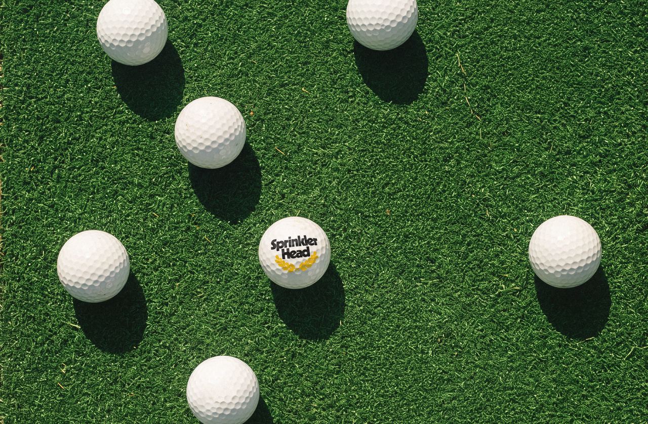 Golf balls scattered on a putting green, one printed with the Sprinkler Head logo